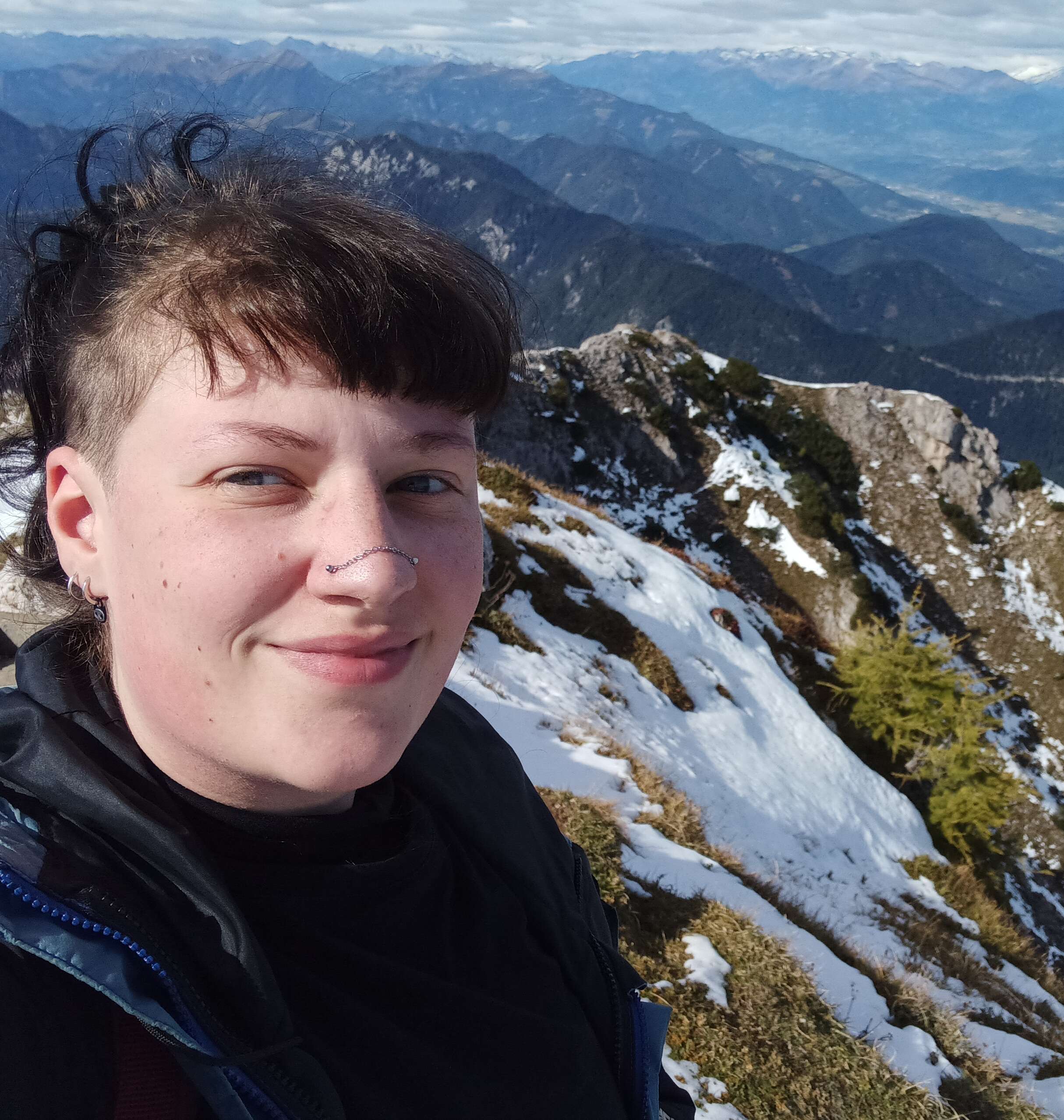 A photo of me at the peak of Dobratsch mountain range. I have pale skin, short black bangs, a chain across my nose, and dark hair tied up to reveal shaved sides.